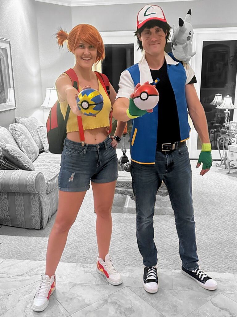 woman in misty from pokemon costume holding a pokeball and with diy misty shoes next to a man in an ash ketchum costume holding a red and white pokeball. pikachu is on the mans shoulder.