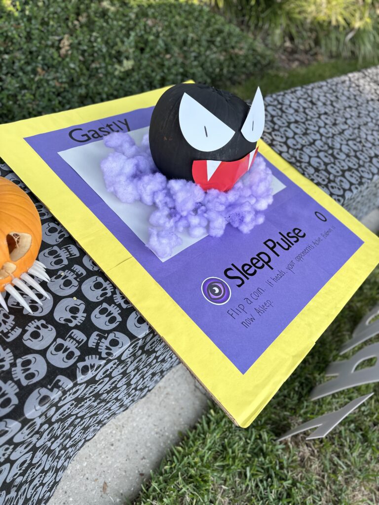 A creatively painted pumpkin transformed into the Pokémon character Gastly, displayed on a vibrant poster with information about its “Sleep Pulse” ability. The pumpkin features large white and black eyes, a red mouth, and is surrounded by purple cloud-like forms to mimic Gastly’s ghostly appearance. The background includes a tablecloth with skull patterns, adding to the Halloween theme. One of the most creative pumpkin painting ideas.