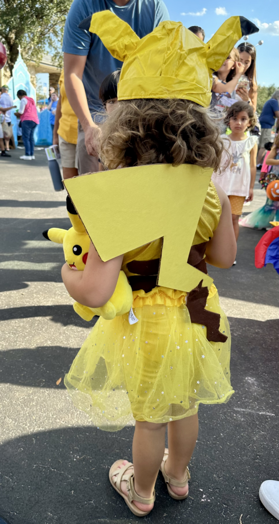 Child in a DIY Pikachu costume from a Pokémon Family Costumes theme, featuring a yellow tutu, handmade hat with black-tipped ears, and a cardboard tail, standing at a Trunk or Treat event.