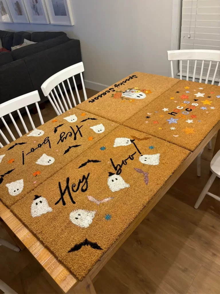 DIY Halloween doormat painting party with four hand-painted doormats featuring ghost designs, 'Spooky Season' text, hocus pocus, and bats on a wooden table indoors.