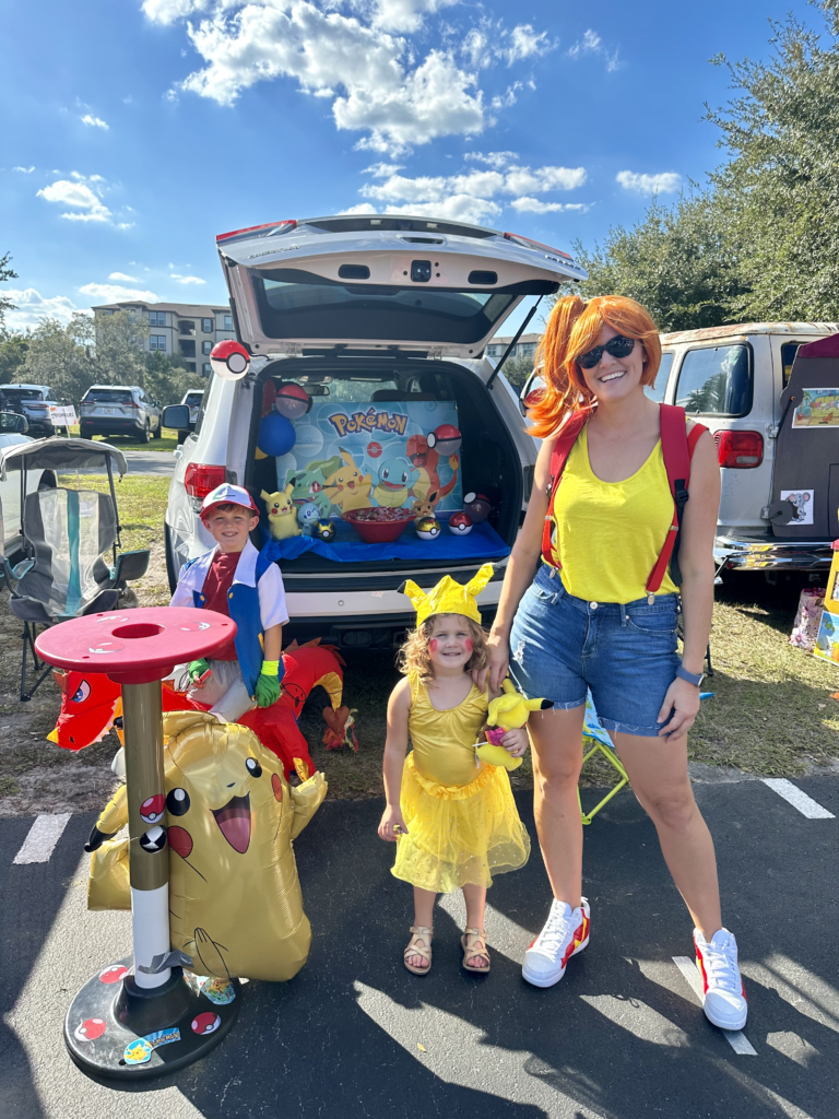 A family dressed in Pokémon-themed costumes for a Trunk or Treat event. The mother is dressed as Misty, the daughter as Pikachu in a yellow tutu and DIY hat, and the son as Ash Ketchum riding a Charizard costume. They are standing in front of their decorated car, which is themed with Pokémon characters and Pokéballs, capturing the festive atmosphere of the event. The scene is bright and colorful, with a clear blue sky and other participants in the background.