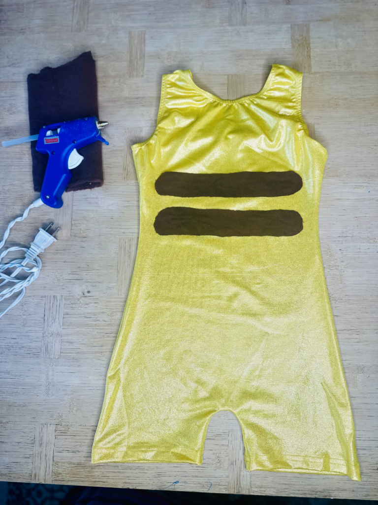 A DIY Pikachu costume in progress, featuring a sparkly yellow leotard with brown felt stripes glued onto the back to mimic Pikachu’s signature look. The leotard is laid out flat on a table next to a hot glue gun and a piece of brown felt, showing the materials and tools used in the crafting process.